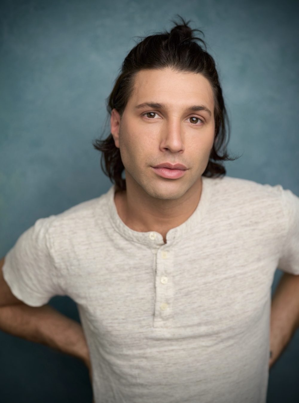 Noah with his hair pulled up, in a cream henley against a blue backdrop