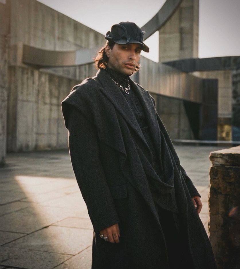 NoahBOY in a draped charcoal coat, black cap and chain, against brutalist concrete